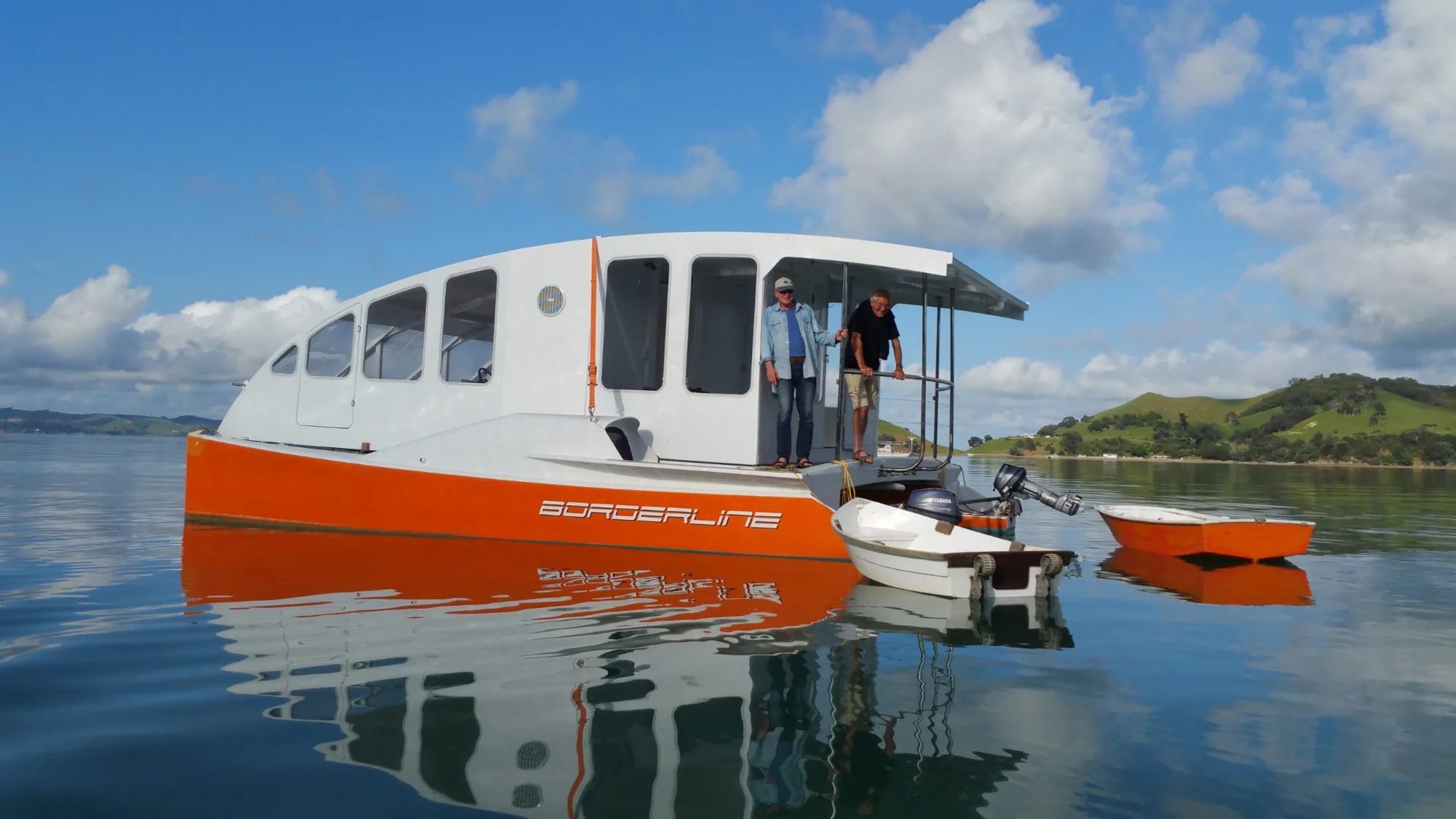 White-cabin catamaran houseboat with orange hulls on calm water, two people aboard