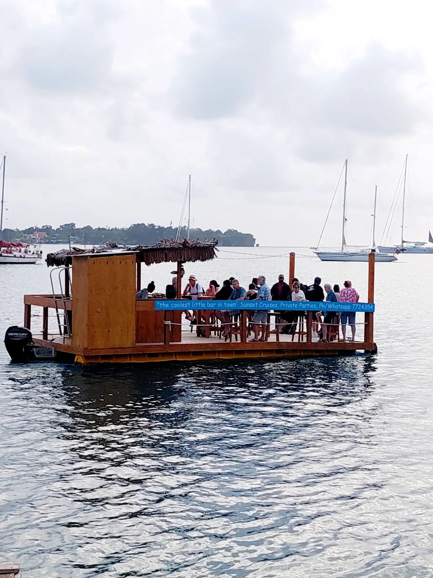 Timber tiki bar boat full of guests on a harbour, yachts behind