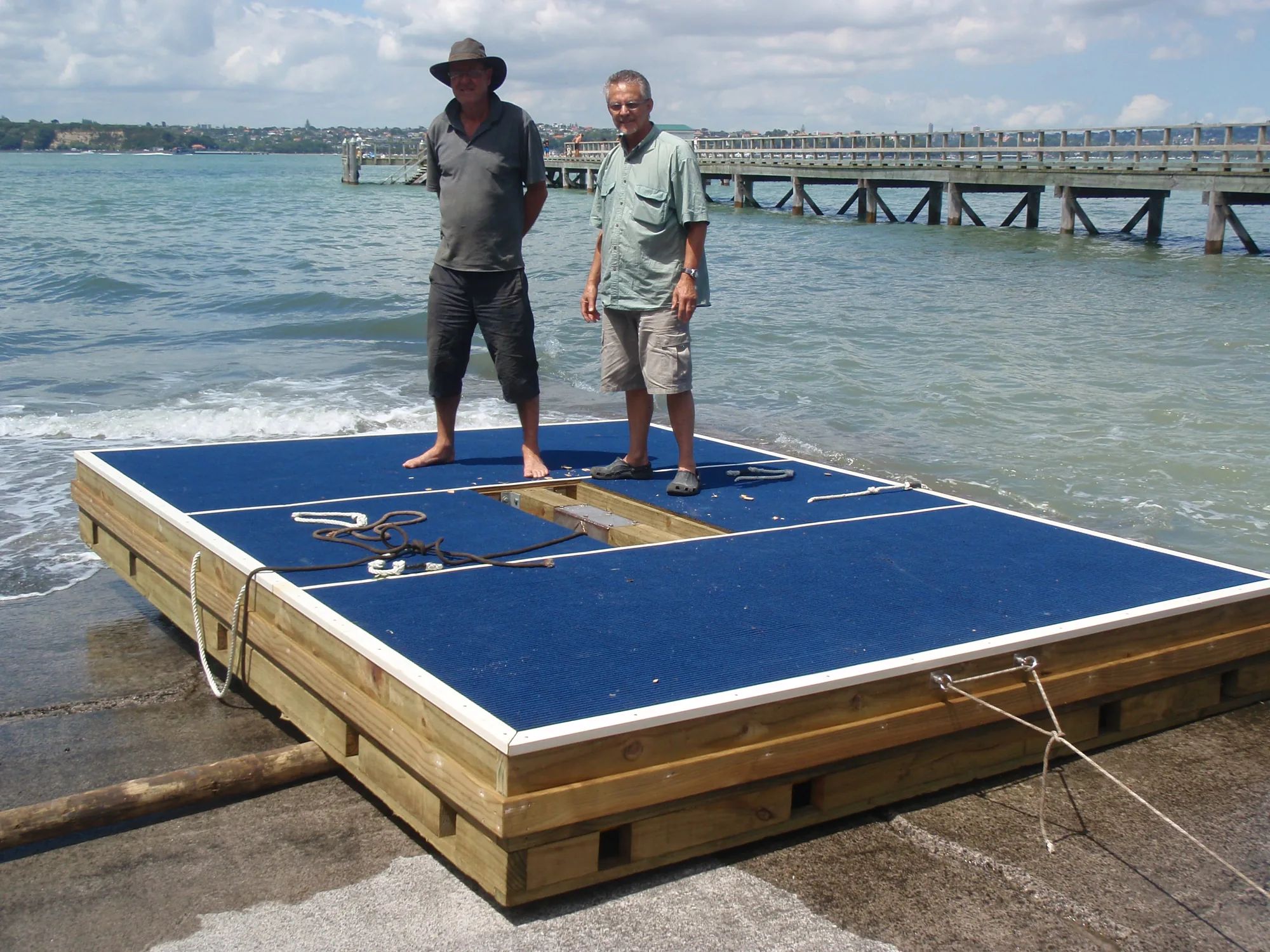 Two men standing on a large blue-decked raft at a boat ramp