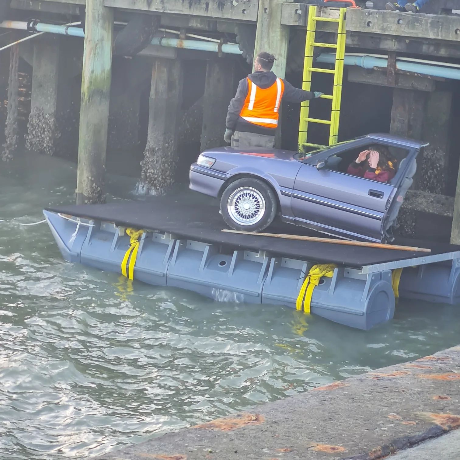 Car on a black-decked pod barge beside wharf piles, crew member standing by