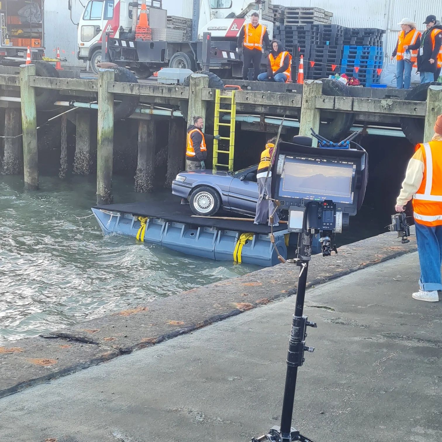 Film crew on a wharf beside a pod barge carrying a car, camera monitor in front