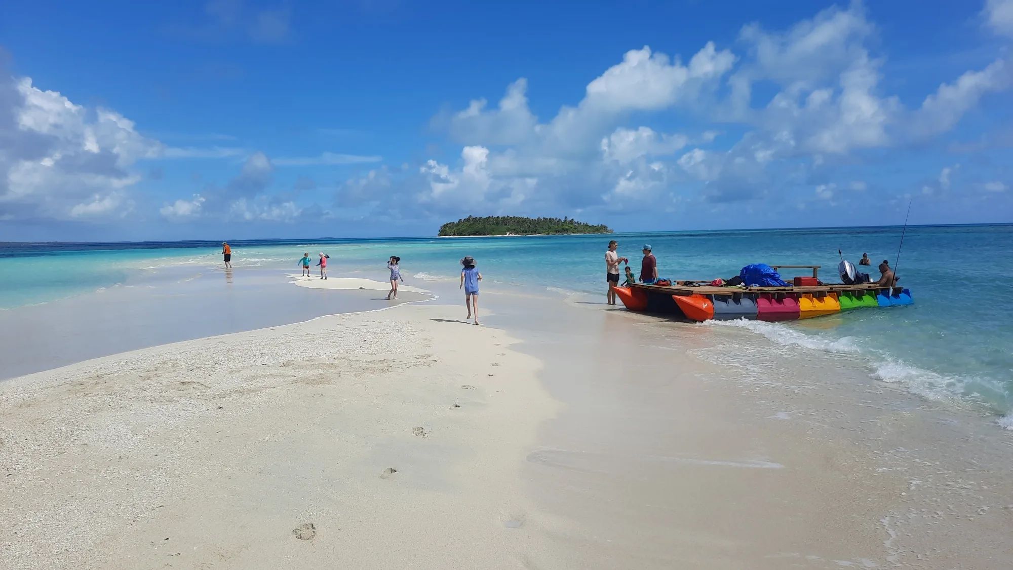 Boat on multicoloured pods at a sandbar, with an island behind and children on the sand