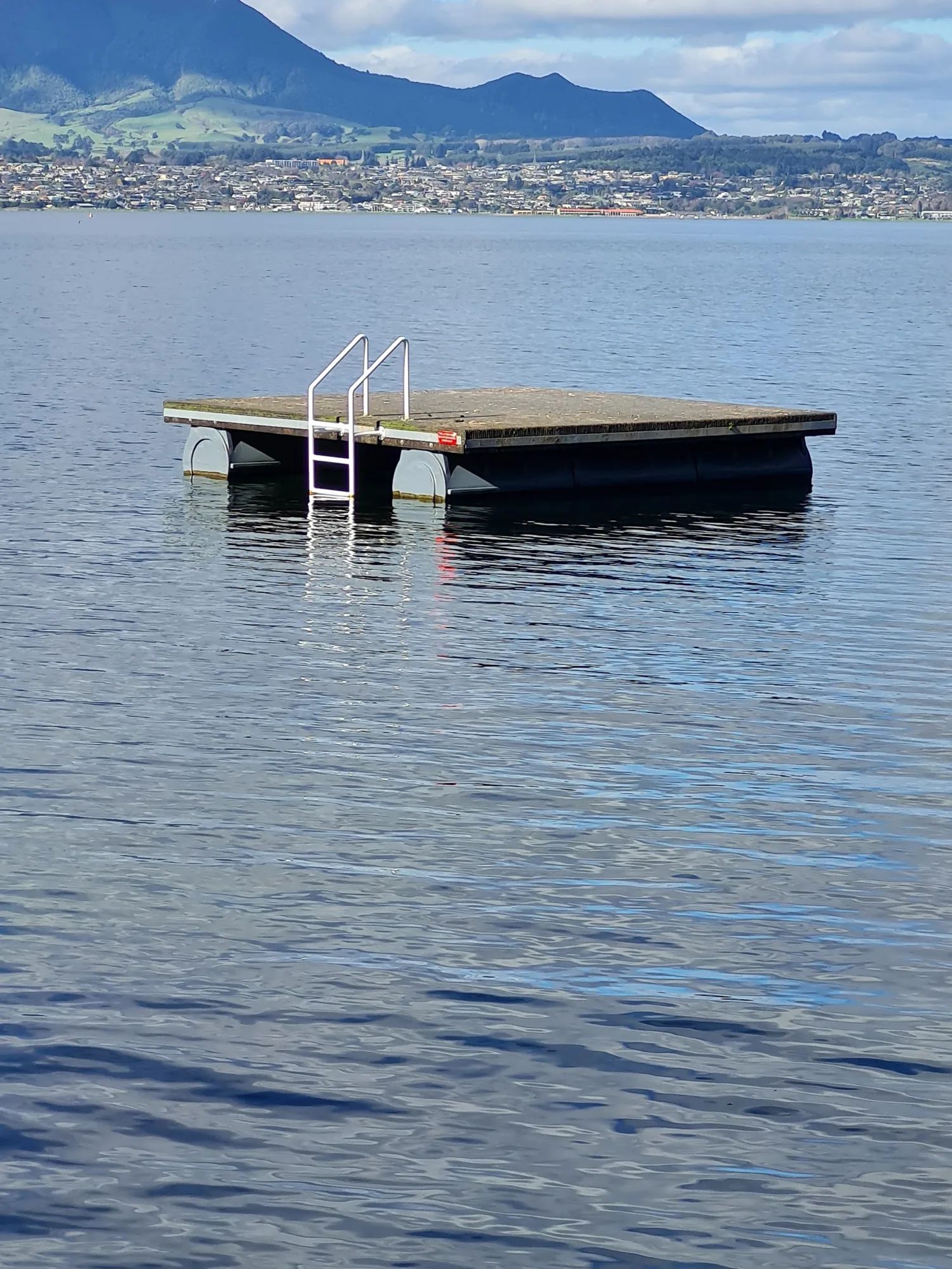 Swim raft with a ladder anchored on a calm lake, town and hills behind